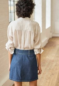 Vaalean beige nappipaita, jossa on käännetyt hihat, yhdistetty tummansinisempään A-linjaisiin hameeseen, jossa on ommeltu vyötärö ja taskuyksityiskohtia.