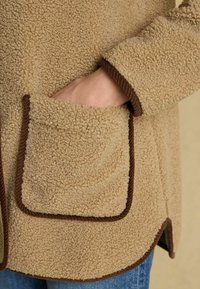 Karamelli fliisjakk, millel on tekstureeritud välimus, pruun äärestus ja esikott. Disain sisaldab ümarat äärt ja nähtavaid õmblustead.