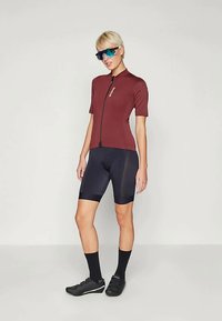 Czerwono-bordowa koszulka rowerowa z krótkim rękawem i zamkiem z przodu, w połączeniu z czarnymi spodenkami z wkładką. Model nosi okulary przeciwsłoneczne i czarne buty rowerowe.