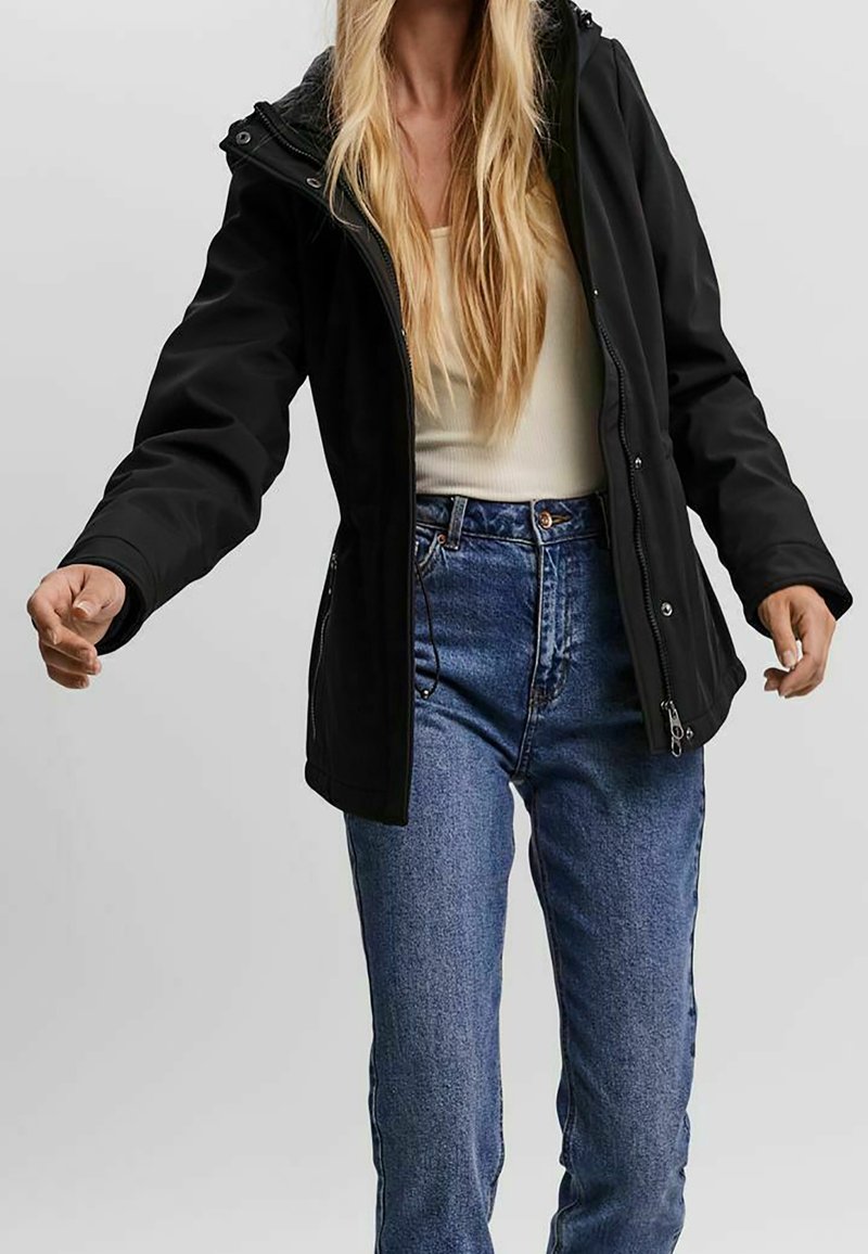 Femme portant une veste noire zippée par-dessus un haut blanc et un jean bleu, debout les bras légèrement étendus sur un fond uni.