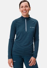 Pull-over long à manches, couleur teal foncé, avec un col haut et un zip mi-hauteur ; présente un logo sur la poitrine et un tissu texturé. Associé à un pantalon assorti.