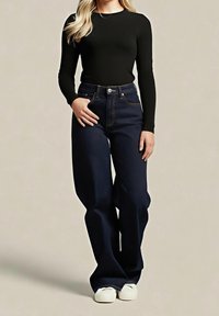 Femme portant un haut noir à manches longues, un jean bleu foncé taille haute et jambes larges et des chaussures blanches, debout avec une main dans la poche.