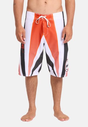 Homme portant un short de bain mi-long avec des motifs géométriques orange, blanc et noir, debout pieds nus sur un fond uni.