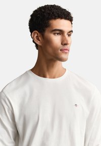 Άνδρας με κοντά κατσαρά μαλλιά, που φορά ένα απλό λευκό μακρυμάνικο πουκάμισο, κοιτάζοντας ελαφρώς προς τα δεξιά σε ουδέτερο φόντο.