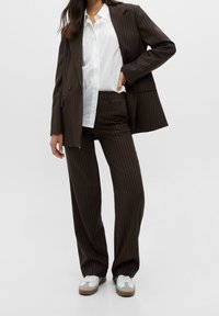 Brunrandig kostym med en skräddarsydd kavaj och lösa byxor. Bärs över en vit skjorta med knappar och kompletteras med vita sneakers.