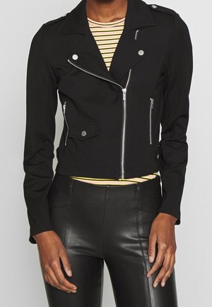 Femme portant une veste courte de motard noire avec des zips argentés par-dessus une chemise rayée jaune et blanche et un pantalon en cuir noir.