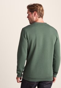 Man with light brown hair wearing a plain green sweatshirt and dark jeans, standing facing away against a beige background.