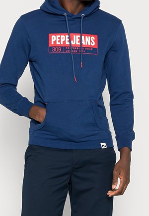 Homme portant un sweat à capuche bleu avec le texte "PEPE JEANS 309 Portobello Road London 1973" et un pantalon bleu assorti, main dans la poche avant.