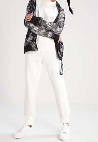 Veste à motifs noir et blanc portée sur un sweat-shirt blanc et un pantalon de survêtement crème ; assortis à des baskets blanches avec une étiquette sur le pantalon.