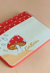 Tarjetero rojo con lunares sobre una superficie crema que presenta una ilustración de un hongo rojo y "Cath Kidston" grabado en dorado. Textura suave.