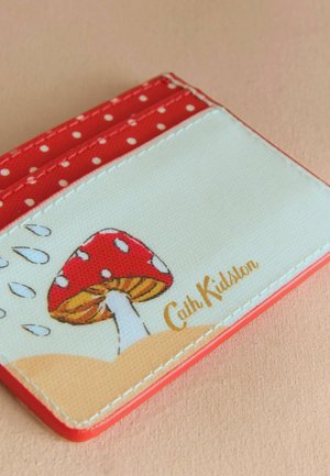 Červený pouzdro na karty s puntíky a krémovým povrchem, na kterém je ilustrace červené houbě a zlato embosované "Cath Kidston". Hladký povrch.