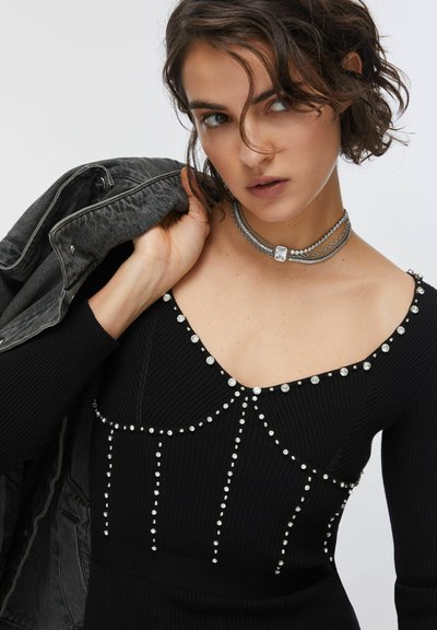 Robe noire côtelée avec des clous argentés, un décolleté carré et une coupe ajustée. Le modèle tient une veste en denim grise sur l'épaule.