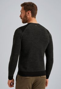 Man met een baard, gekleed in een donkergrijze gestructureerde trui en een paarse broek, met zijn rug naar de camera tegen een effen lichtgrijze achtergrond.
