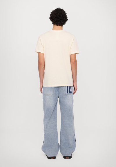 Cremfarbene T-Shirt mit kurzen Ärmeln, kombiniert mit hellblauen, weit geschnittenen Jeans mit seitlichen Streifen und strukturiertem Finish, von hinten betrachtet.
