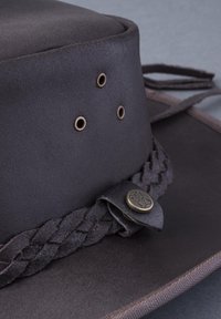 Brown leather hat with a smooth texture, featuring three ventilation grommets, a braided band, and a decorative metal button detail.