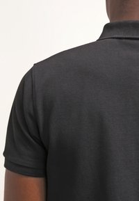 Personne portant un polo noir à manches courtes vue de dos sur un fond clair uni.
