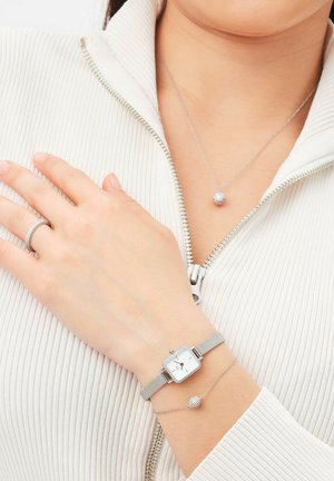 Strieborné pletivo hodinky so štvorcovým ciferníkom, akcenty s kryštálmi; jemný strieborný náhrdelník s guľovým príveskom; pruhovaný biely zipsový top.