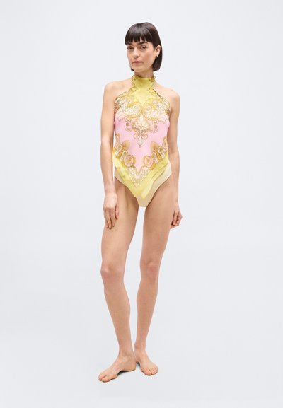 Γυναίκα που στέκεται ξυπόλυτη φορώντας ένα κίτρινο και ροζ κορμάκι με λουράκι στον λαιμό, διακοσμημένο με περίτεχνα χρυσά μοτίβα, μπροστά σε απλό φόντο.