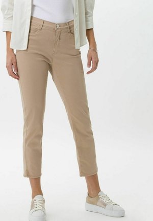 Femme portant un pantalon beige court, des baskets blanches, un débardeur blanc et une chemise blanche déboutonnée, debout devant un fond uni.