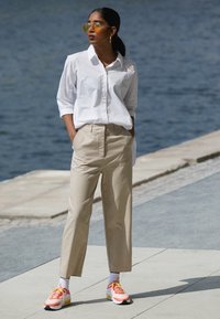 Vit knäpp skjorta, beige skräddarsydda byxor, klarröda och gula sneakers. Modellen bär överdimensionerade gula solglasögon och står vid vattnet.