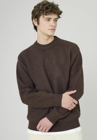 Pull en tricot brun avec un col rond, des poignets et un ourlet côtelés ; texture douce ; coupe décontractée ; porté par-dessus un t-shirt blanc.