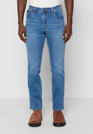 Mężczyzna w średniobłękitnych prostych dżinsach i brązowych zamszowych butach, stoi na jednolitym jasnoszarym tle.