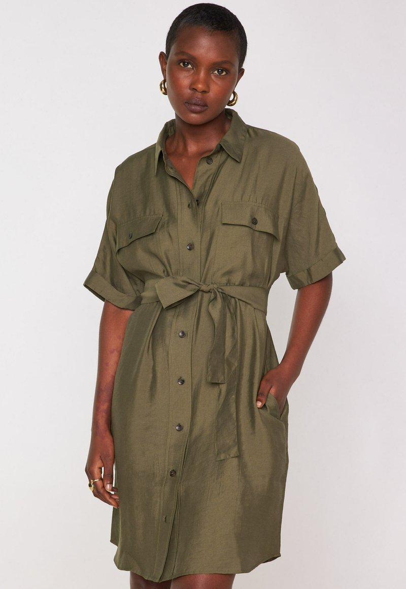 Abito camicia verde oliva con maniche corte, chiusura a bottoni, vita con cintura e due tasche sul petto. Tessuto morbido con una vestibilità rilassata.