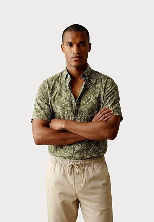 Mann mit verschränkten Armen, trägt ein kurzärmeliges grünes Hemd mit Blattmuster und beigefarbene Hose mit Kordel, steht vor einem einfarbigen hellen Hintergrund.