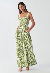 Robe maxi verte avec des rayures blanches en forme de zèbre, décolleté carré, taille smockée et poches. Fabriquée en tissu léger. Sandales plates portées.