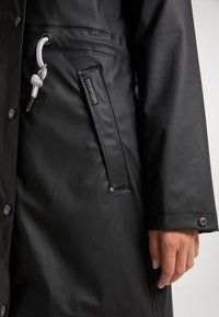 Manteau noir imperméable à la texture lisse, avec une poche latérale, des éléments métalliques argentés et un cordon blanc en accent.