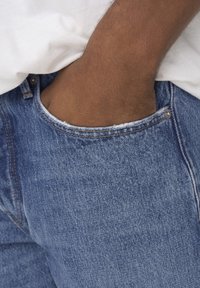 Jeans de mezclilla azul claro con un borde de bolsillo deshilachado y costuras visibles, usados con una camisa blanca. Mano metida en el bolsillo.