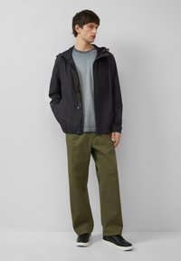 Zwarte lichtgewicht jas met capuchon, gecombineerd met een grijs T-shirt, olijfgroene broek en zwarte schoenen. Eenvoudig ontwerp, relaxte pasvorm.