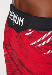 Shorts d'entraînement rouges avec une taille noire arborant le logo "Venum". Le tissu présente des motifs dynamiques dans diverses teintes de rouge et des accents blancs.