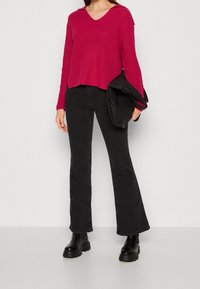 Femme portant un pull en maille rouge, un jean noir évasé, des bottes noires épaisses, tenant une grande pochette noire sur un fond uni.