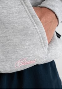 Felpa grigia con una texture morbida, caratterizzata da un logo ricamato rosa sul bordo inferiore e una tasca laterale. Il design include polsini e bordo inferiore a costine.