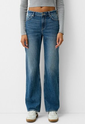 Personne portant un jean bleu taille haute et coupe large, des baskets blanches avec le swoosh noir de Nike, et un haut léger gris clair à manches longues et cropped.