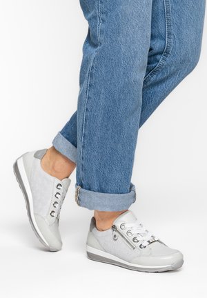 Person trägt hochgekrempelte blaue Jeans und hellgraue Schnürsneaker mit seitlichen Reißverschlüssen, steht auf einer weißen Oberfläche.