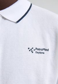 Vit pikétröja i texturerad tyg med marinblå randig krage. Har "Petrol®Ind Daytona"-logotyp i mörkblått.