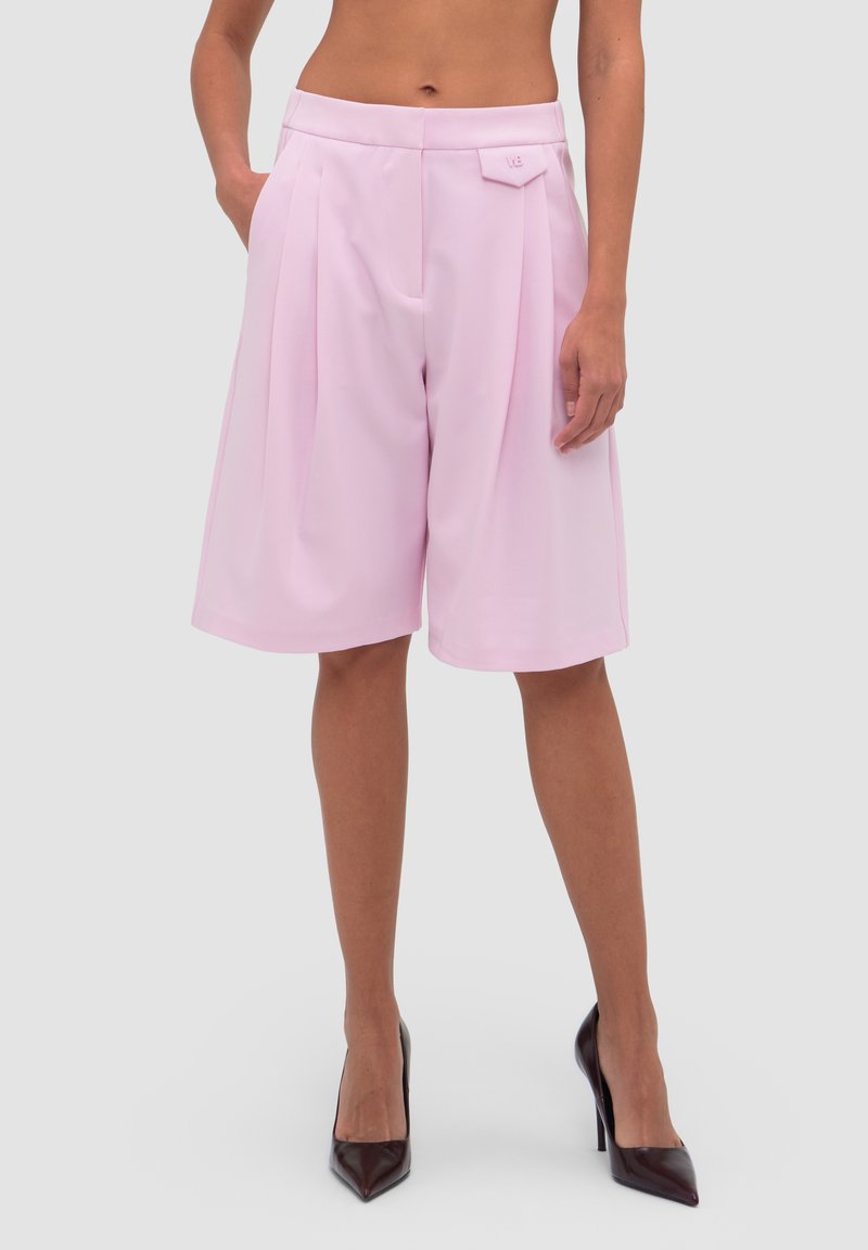 Buste et jambes d'une femme portant un short large taille haute, rose pastel, longueur genou avec plis devant et talons pointus noirs.