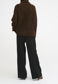 Pull en tricot marron oversize avec un col haut, associé à un pantalon large noir. Le tissu semble doux et texturé.