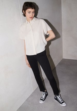 Camisa de manga curta creme com botões, combinada com jeans pretos justos e sapatilhas de cano alto pretas, numa superfície de piso cinza liso.