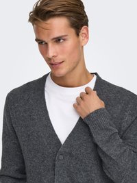 Gray wool cardigan with a V-neck, ribbed cuffs, a single button closure, and a textured knit. Worn over a white T-shirt.
