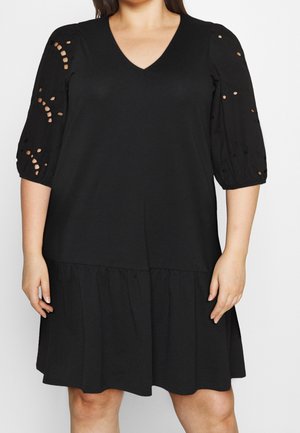 Kobieta ubrana w czarną sukienkę do kolan z dekoltem w serek i bufkami na rękawach o długości trzy czwarte ozdobionymi wzorem dziurek.