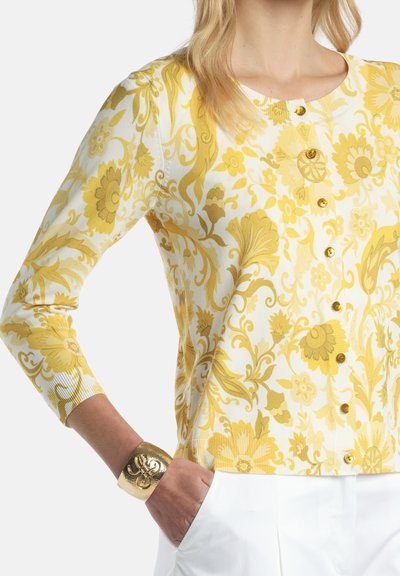 Femme portant un cardigan boutonné à motif floral jaune, un pantalon blanc et un large bracelet en or à son poignet.