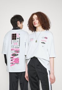 Bílé oversized tričko s různými grafickými potisky v růžové a černé, umístěné na neutrálním pozadí. Nosené obyčejné černé kalhoty.