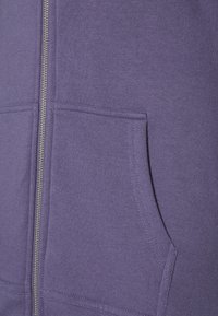 Sudadera con cremallera de color púrpura, hecha de tejido suave. Presenta un bolsillo canguro y detalles acanalados en los puños y el dobladillo. Textura suave.