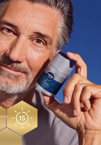 NIVEA Men Age Revitalizing Hyaluron cream in a cylindrical container with a blue label and white body, held by a person against a blue background.