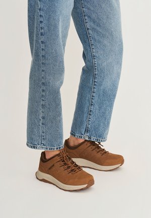 Persoon draagt lichtblauwe spijkerbroek en bruine veter sneakers met dikke witte zolen op een lichte achtergrond.