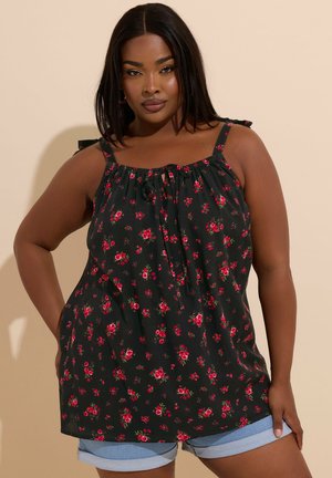 Femme portant un débardeur noir à motifs floraux rouges et un short en jean bleu clair retroussé, debout devant un fond beige.