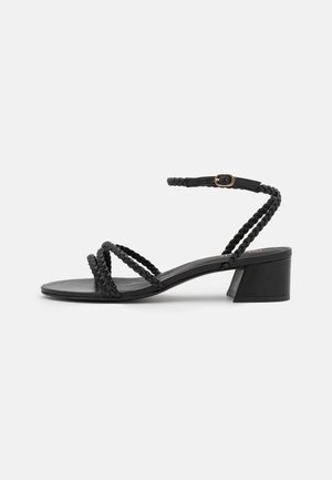 Czarna sandałka na niskim, szerokim obcasie z plecionymi paskami przechodzącymi przez palce i kostkę, zabezpieczona małą złotą klamrą na jednolitym tle.